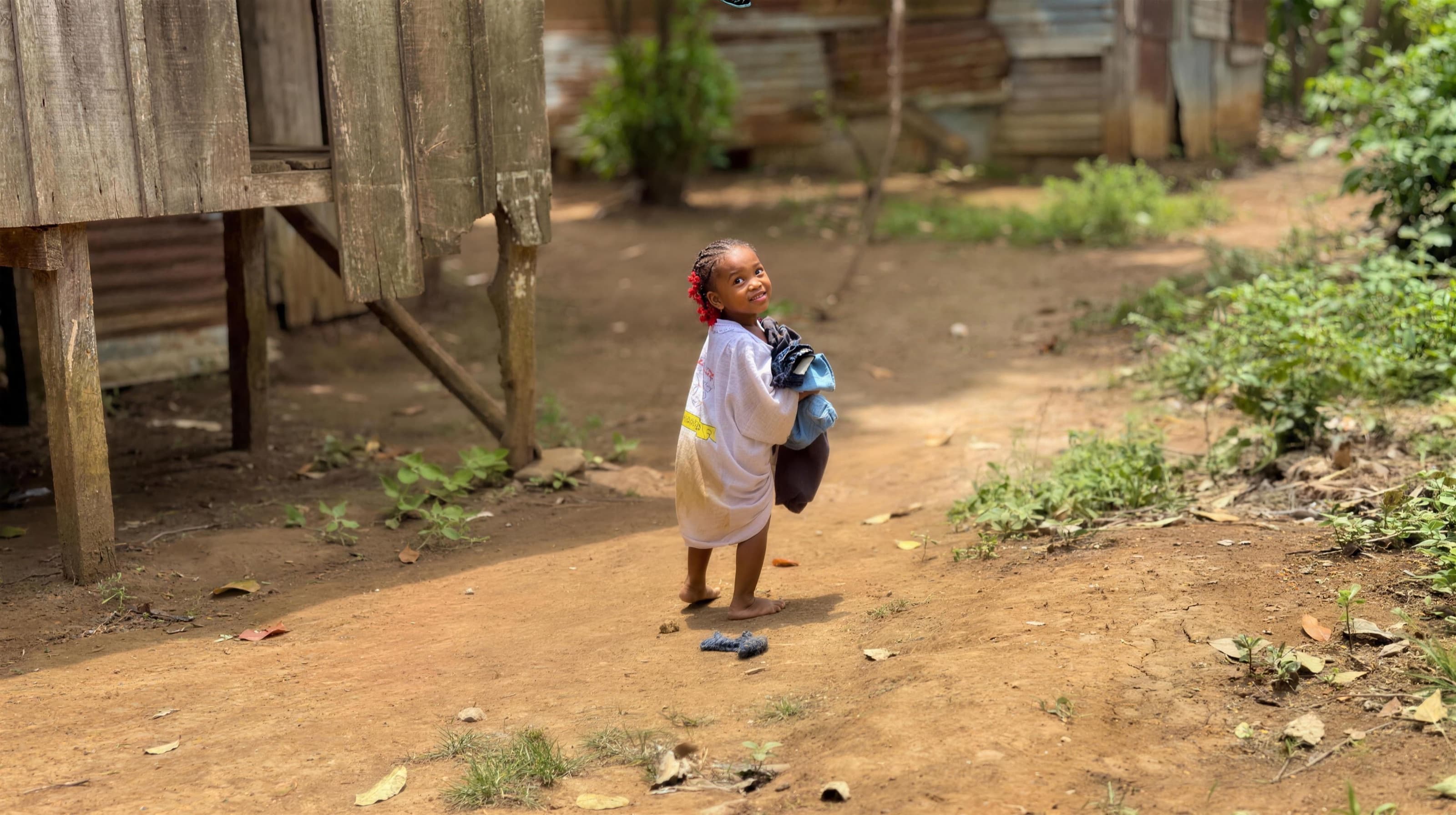 A barefoot girl on a dirt path in a village in São Tomé, holding a bundle of clothes and looking up with a smile.
